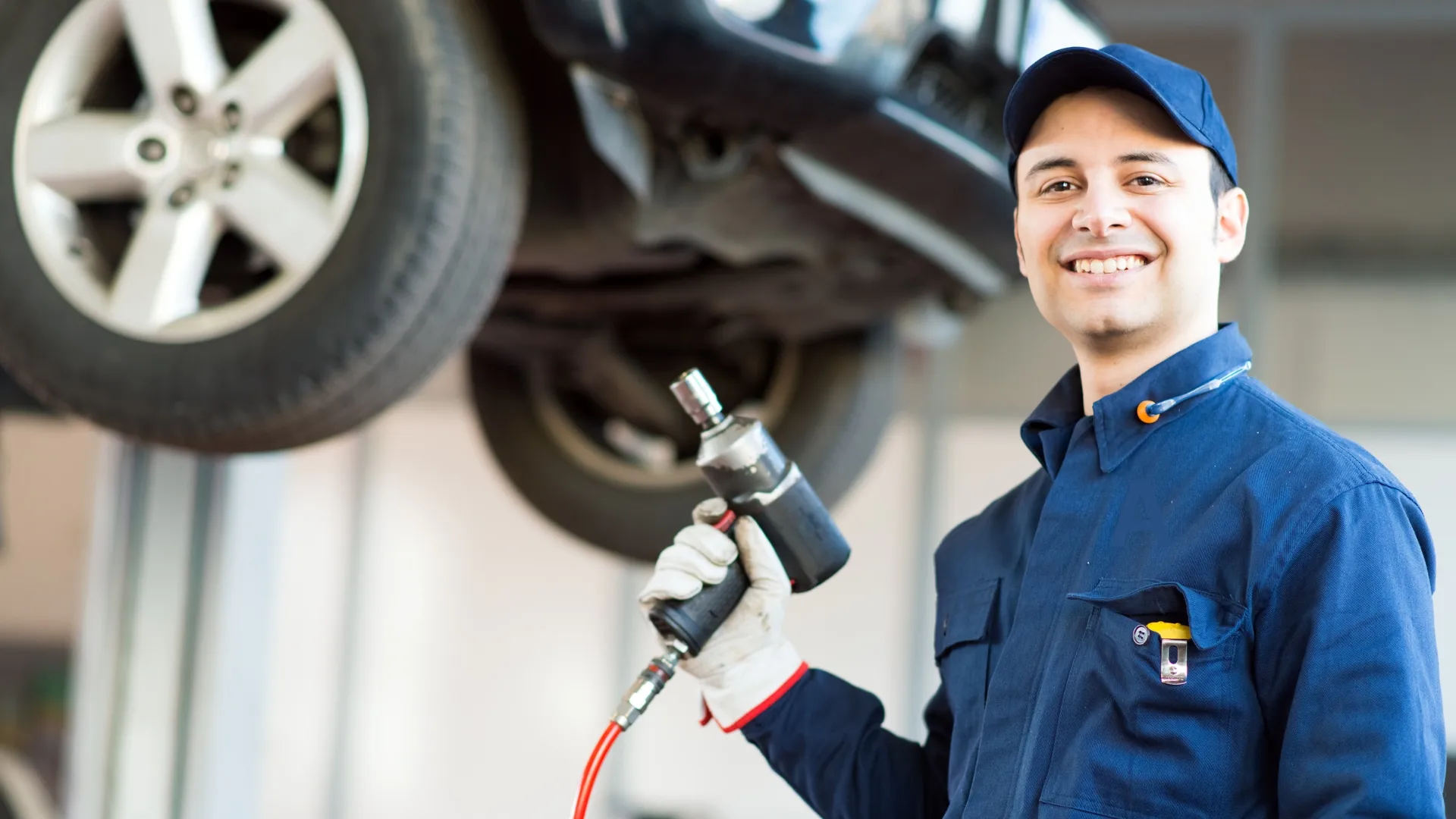 Mechanic in uniform holding a pneumatic wrench with a car on a lift in the background.